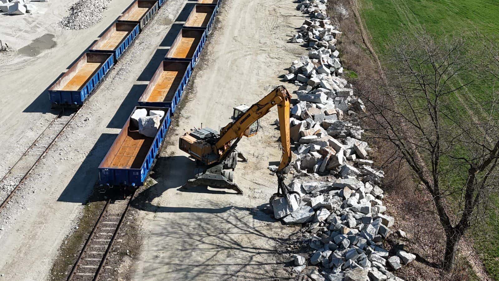 Widok z góry na skład wagonów i koparkę przeładunkową podczas rozładunku kamienia